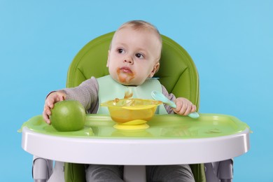 Cute little baby eating healthy food in high chair on light blue background