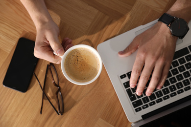 Man with laptop, cup of coffee and smartphone at wooden table, top view