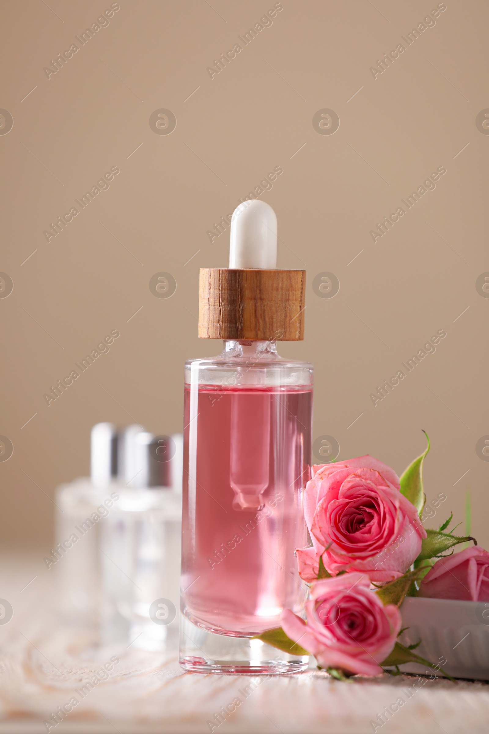 Photo of Bottle of essential rose oil and flowers on white wooden table against beige background