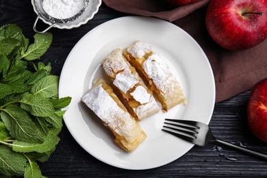 Delicious strudel, powdered sugar, mint and fresh apples on black wooden table, flat lay