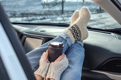 Photo of Young woman in warm socks holding her legs on car dashboard and drinking coffee. Cozy atmosphere