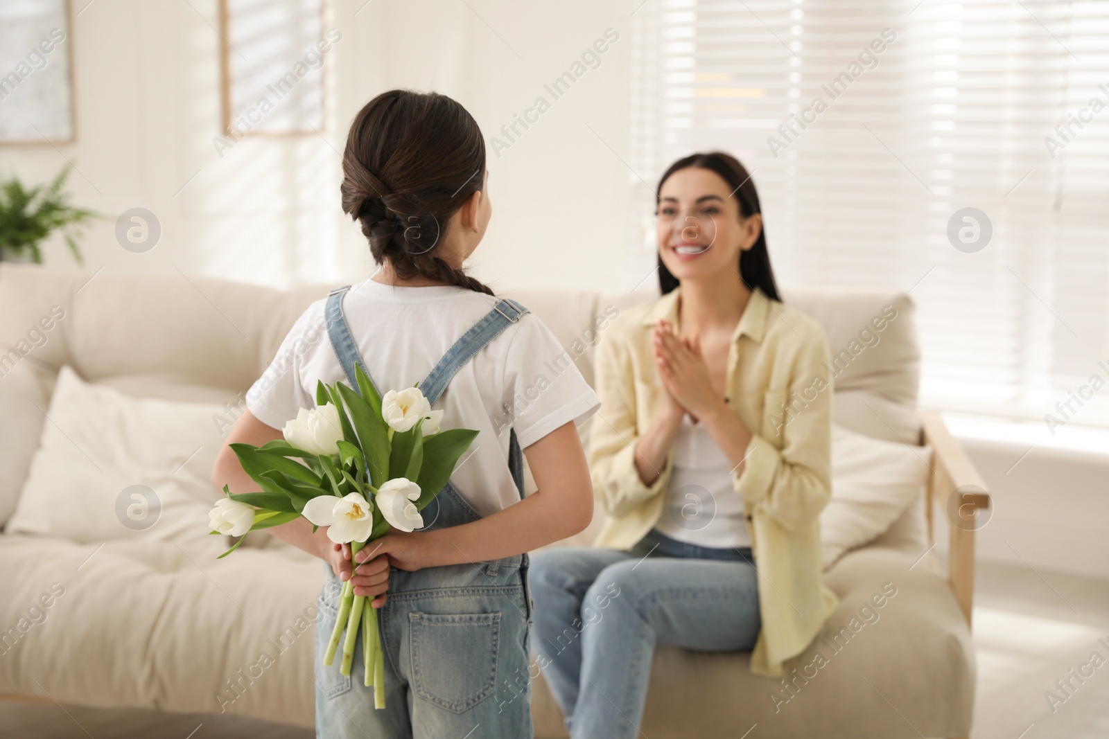 Photo of Little girl hiding tulip bouquet for mom at home. Happy Mother's Day