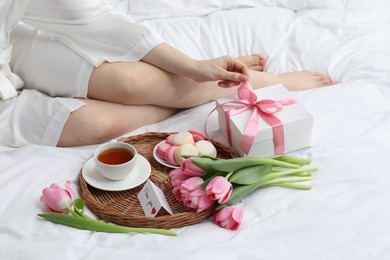 Photo of Tasty breakfast served in bed. Woman with gift box, tea, macarons, flowers and I Love You card at home, closeup