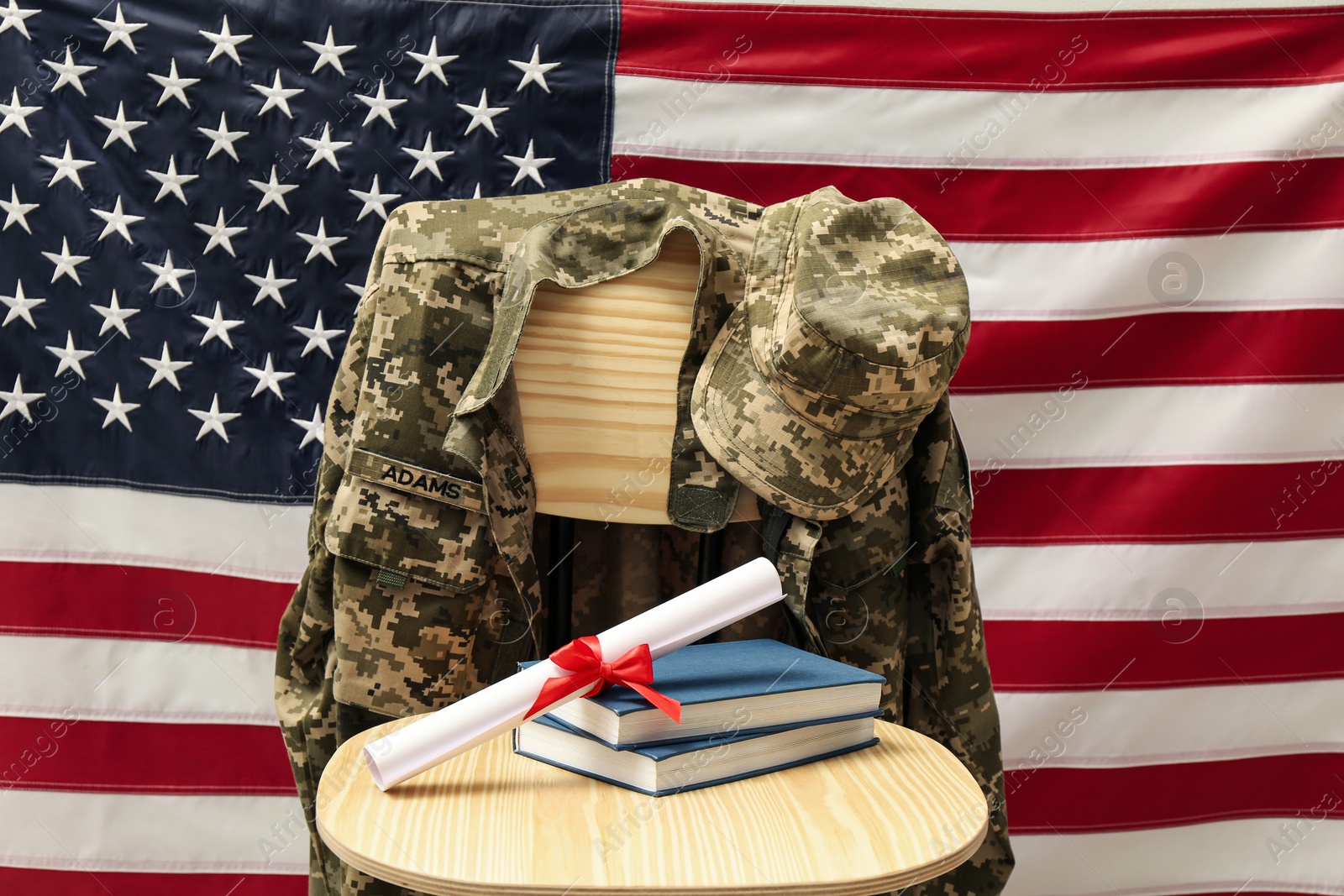 Photo of Diploma, books and soldier uniform on wooden chair near flag of United States. Military education