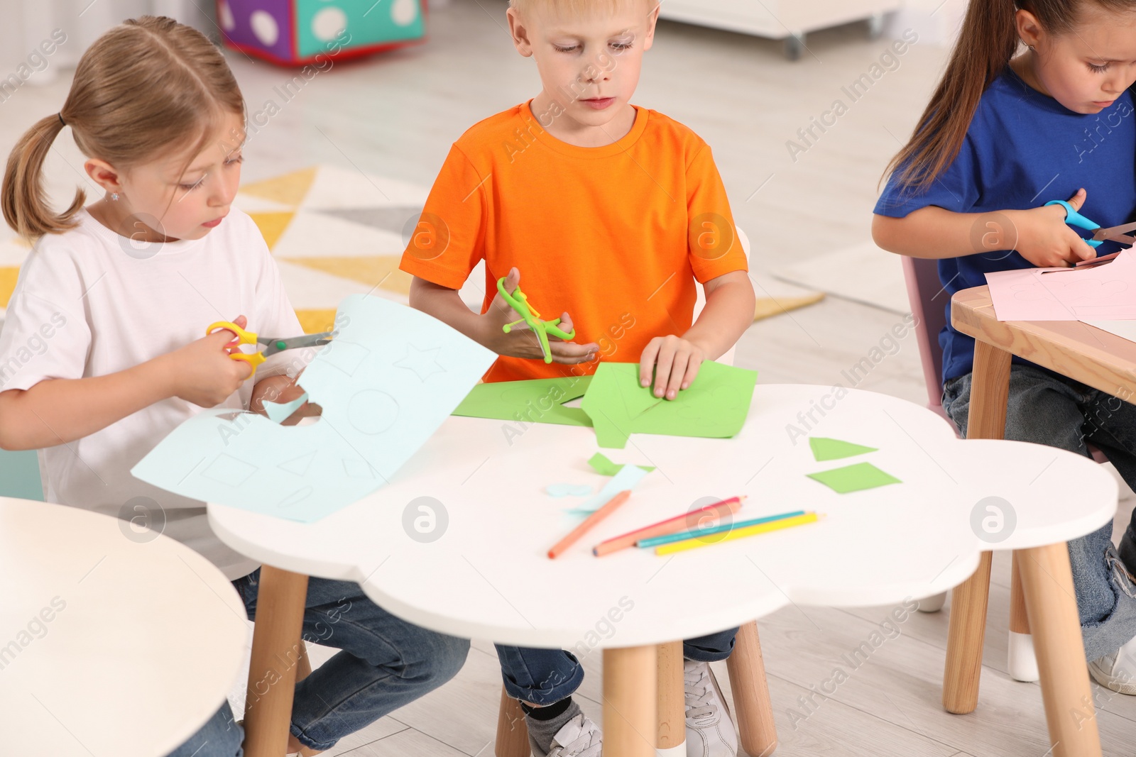 Photo of Cute little children cutting color paper with scissors at desks in kindergarten. Playtime activities