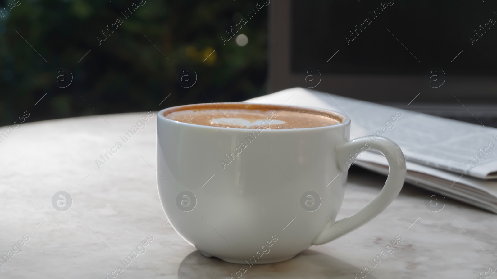Photo of Cup of delicious coffee, newspaper and laptop on beige marble table outdoors