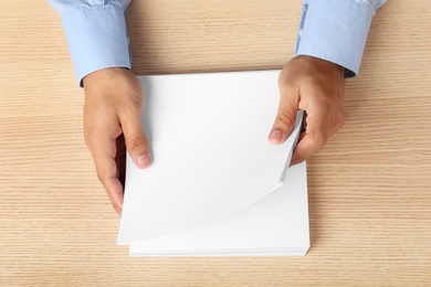 Photo of Man holding blank paper sheets for brochure at wooden table, top view. Mock up