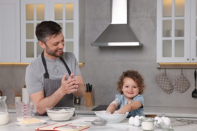 Photo of Cute little girl with her father cooking by recipe book in kitchen