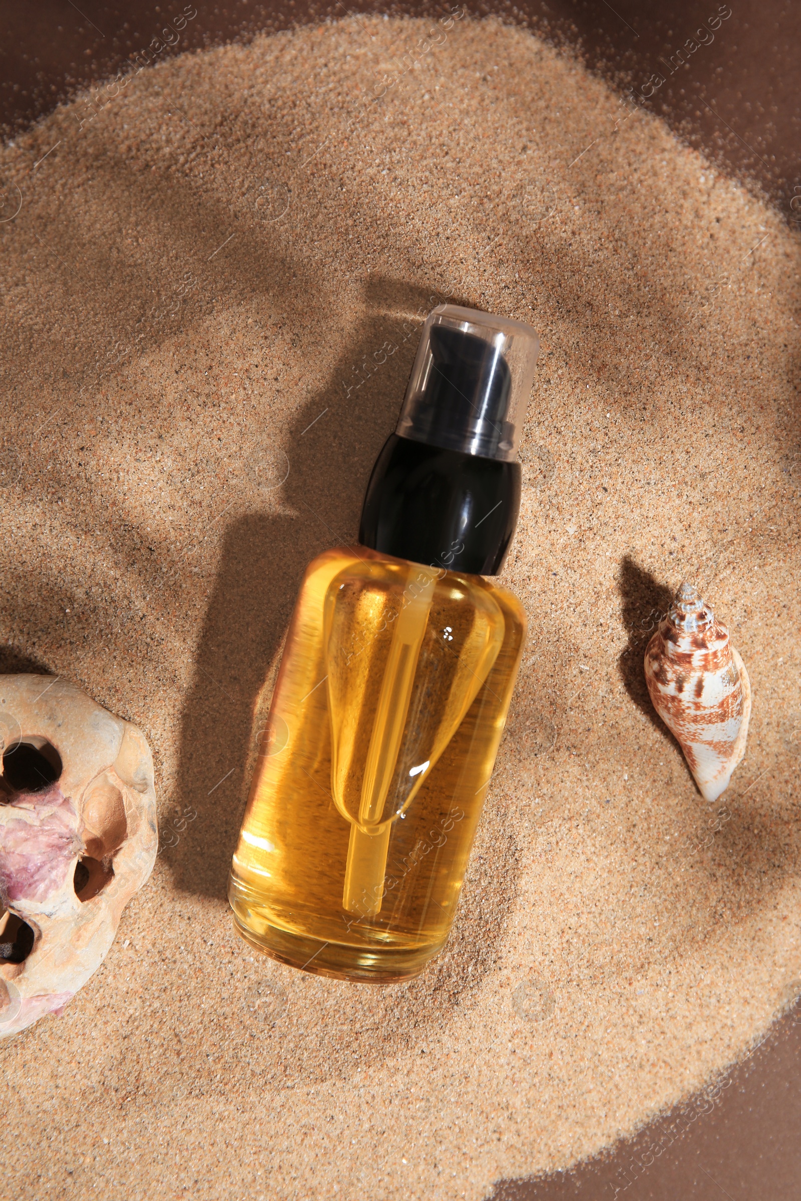 Photo of Bottle of serum, seashell and stone on sand against brown background, flat lay