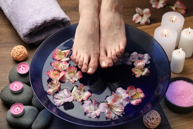 Photo of Woman holding her feet over bowl with water and flowers on wooden floor, closeup. Spa treatment