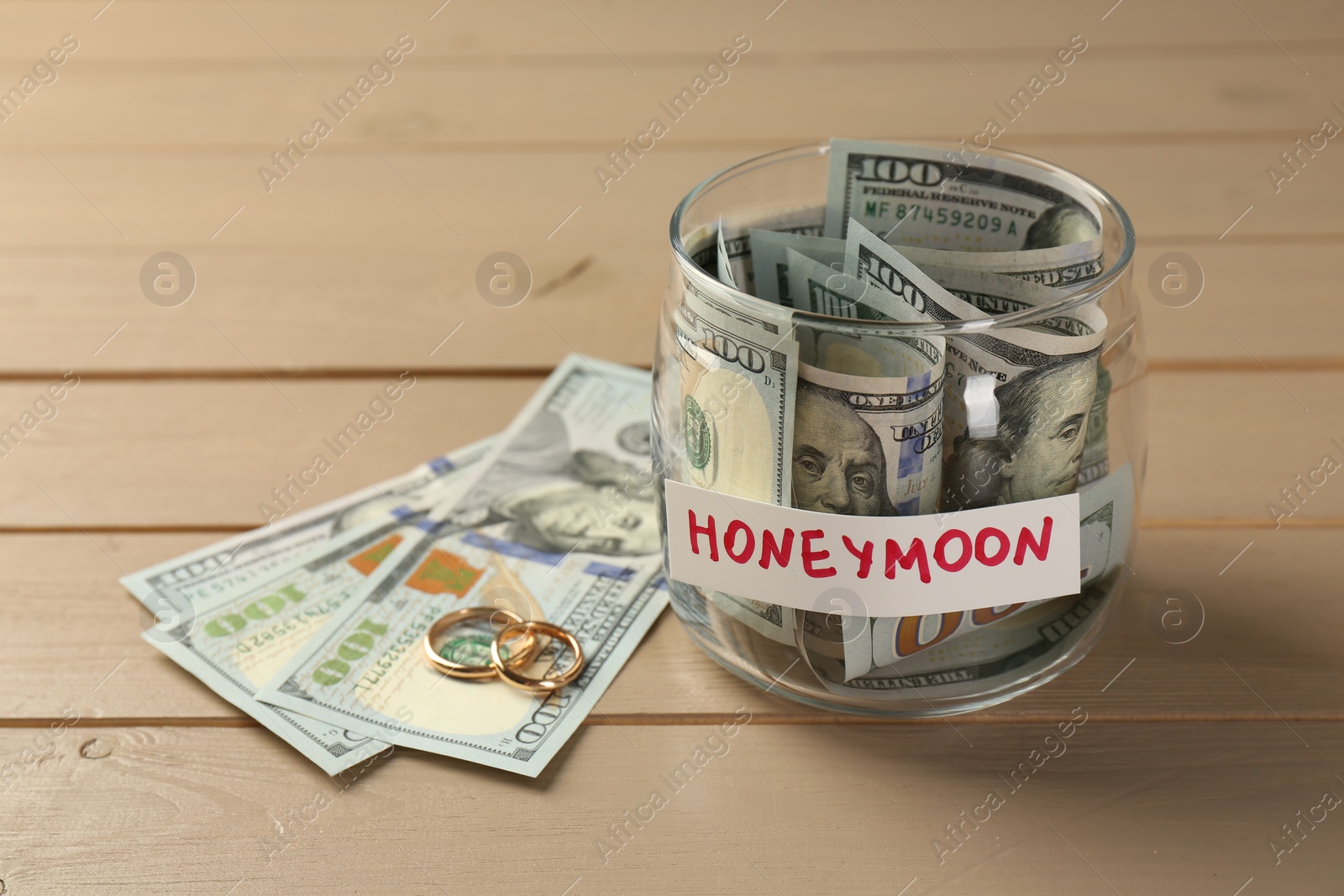 Photo of Glass jar with word Honeymoon, dollar banknotes and golden rings on wooden table, closeup