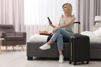 Smiling guest with smartphone and suitcase relaxing on bed in stylish hotel room