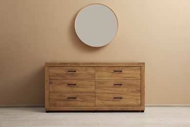Wooden stylish chest of drawers near light brown wall with round mirror