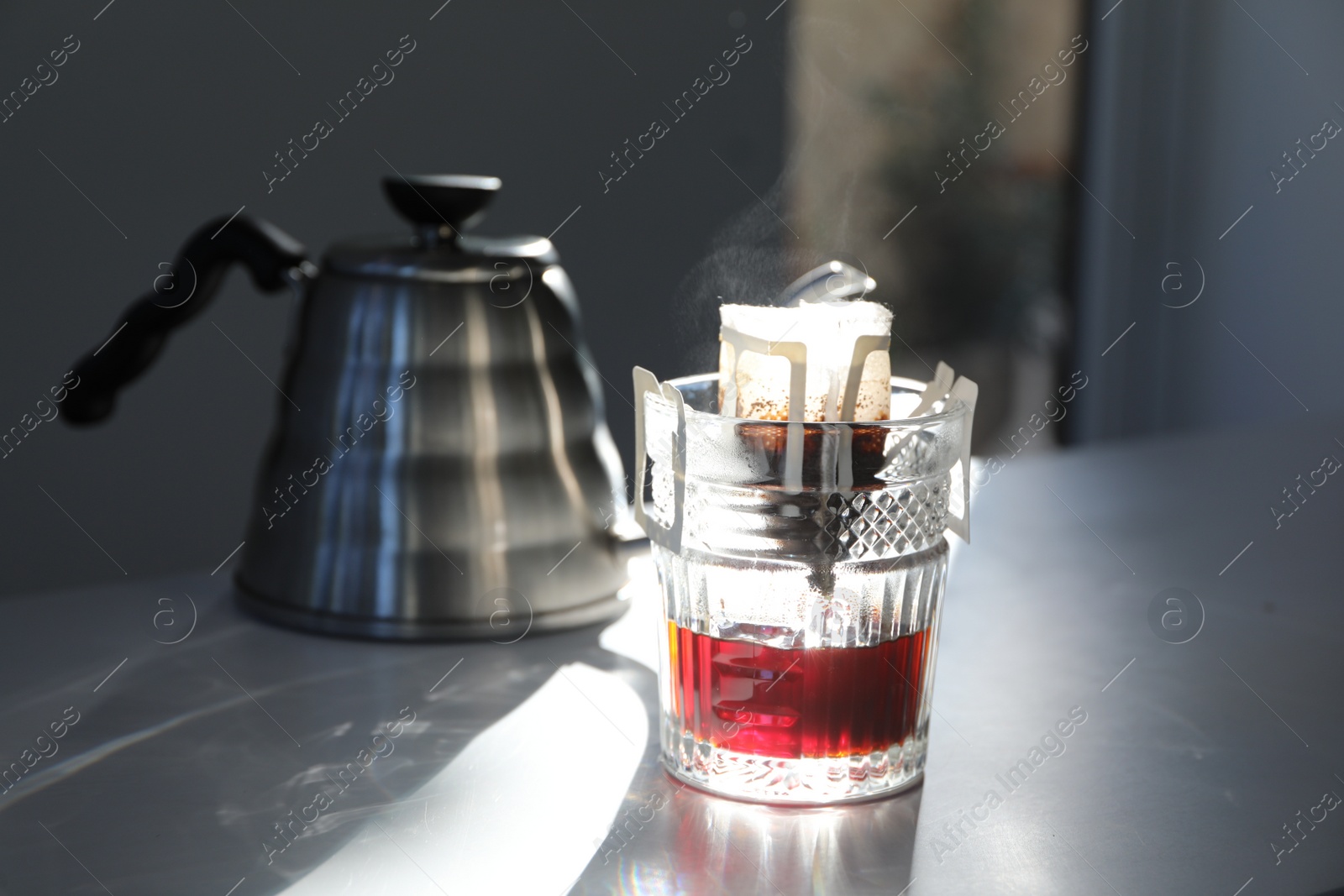 Photo of Glass with drip coffee bag and kettle on light grey table