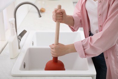 Photo of Woman using plunger to unclog sink drain in kitchen, closeup
