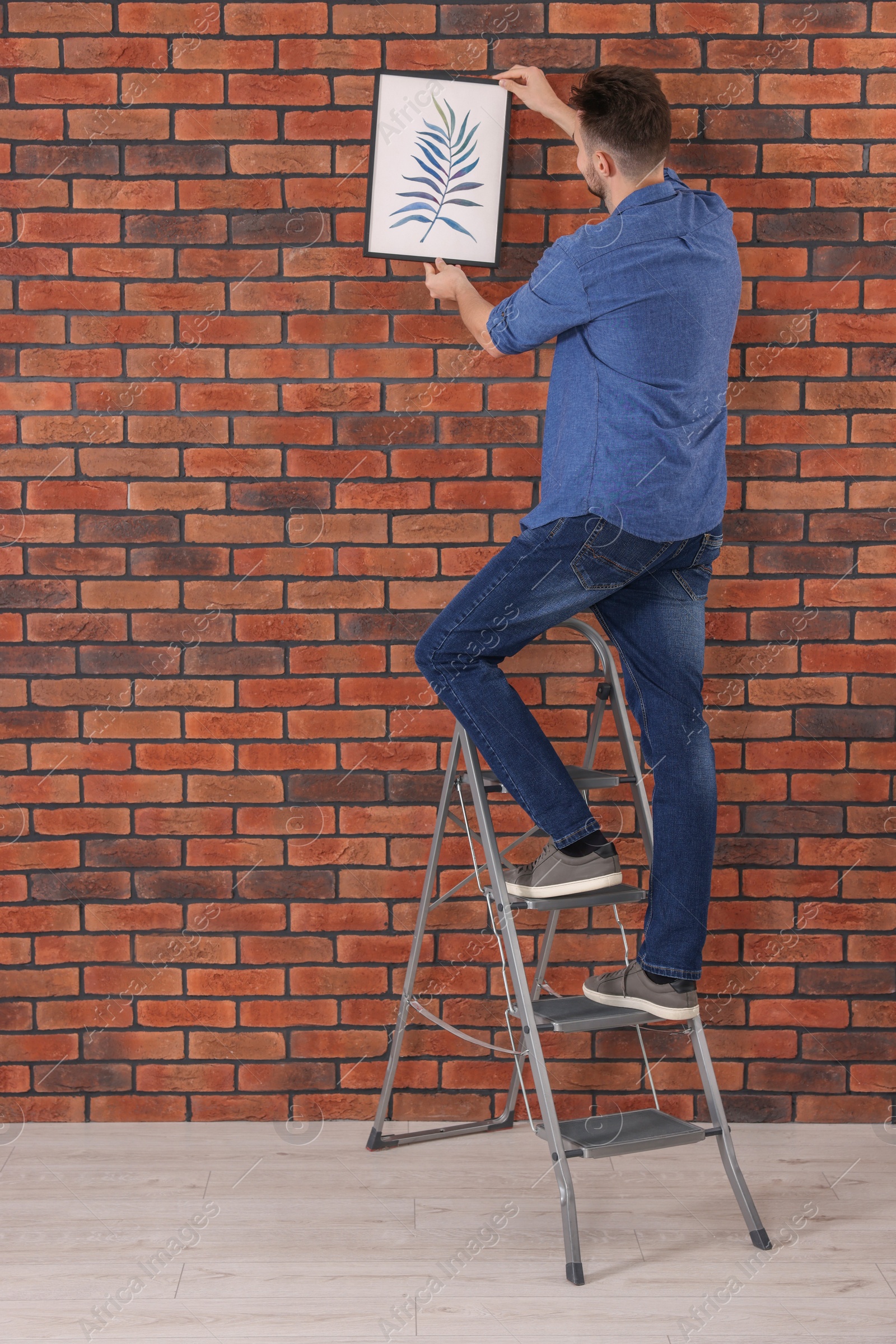 Photo of Man on stepladder hanging picture near red brick wall