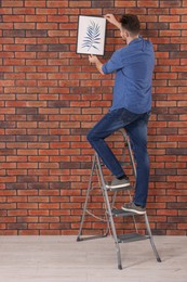 Photo of Man on stepladder hanging picture near red brick wall