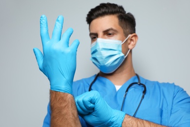 Doctor in protective mask putting on medical gloves against light grey background