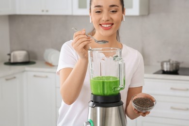 Photo of Beautiful young woman adding chia seeds into delicious smoothie in kitchen, focus on blender