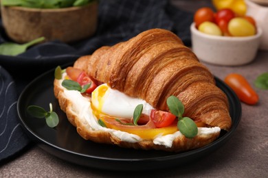 Photo of Tasty croissant with fried egg, tomato and microgreens on brown textured table, closeup