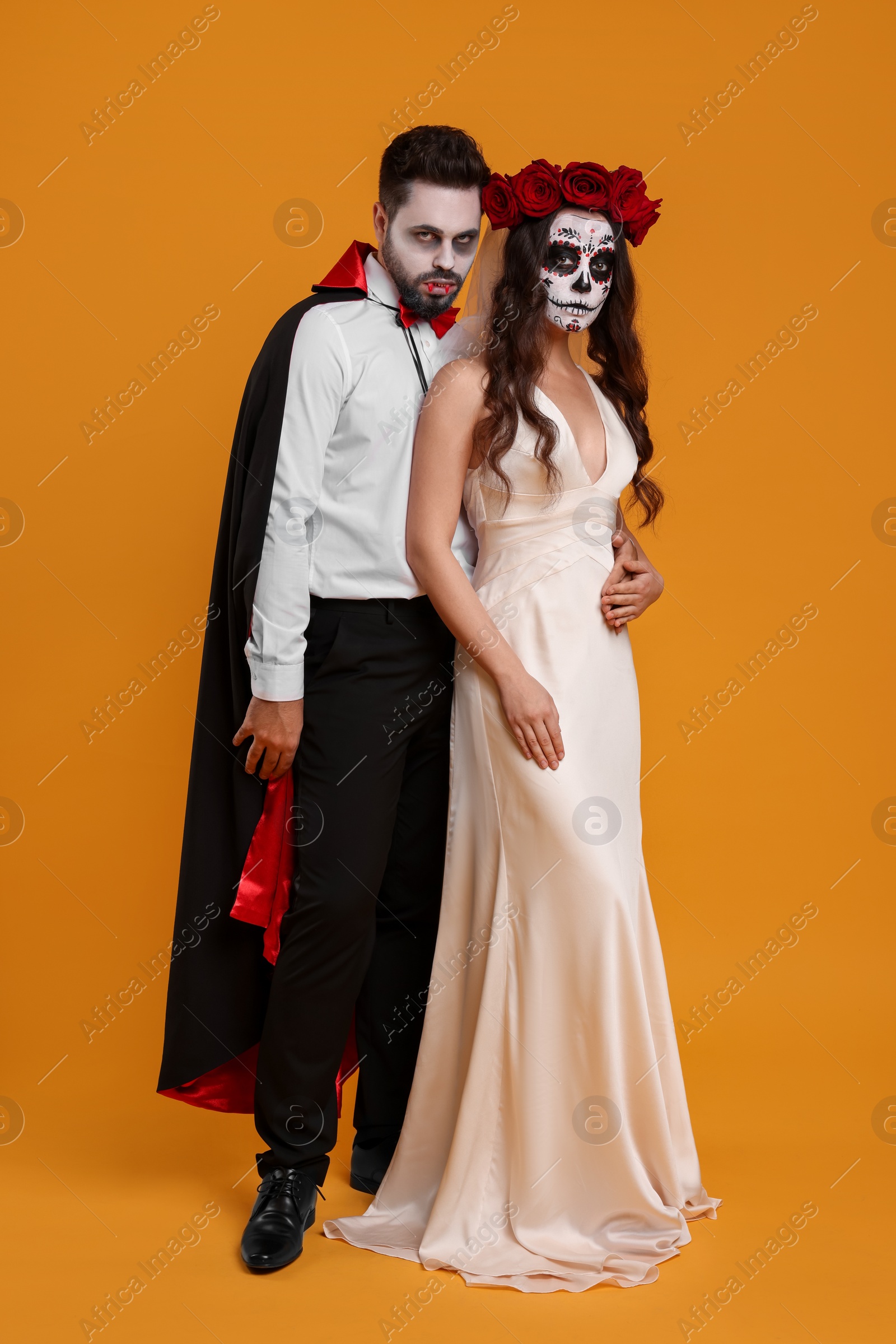 Photo of Couple in scary bride and vampire costumes on orange background. Halloween celebration