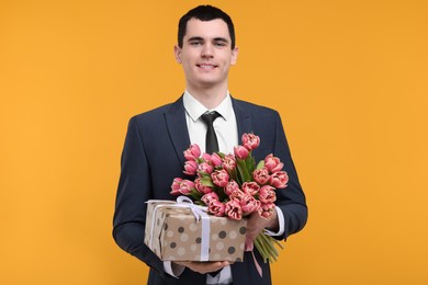 Happy young man with beautiful bouquet and present on orange background