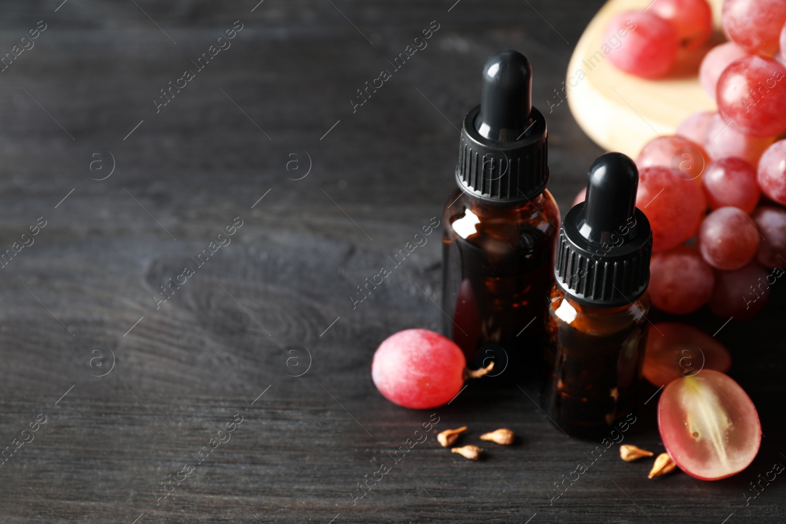 Photo of Organic red grapes, seeds and bottles of natural essential oil on black wooden table. Space for text