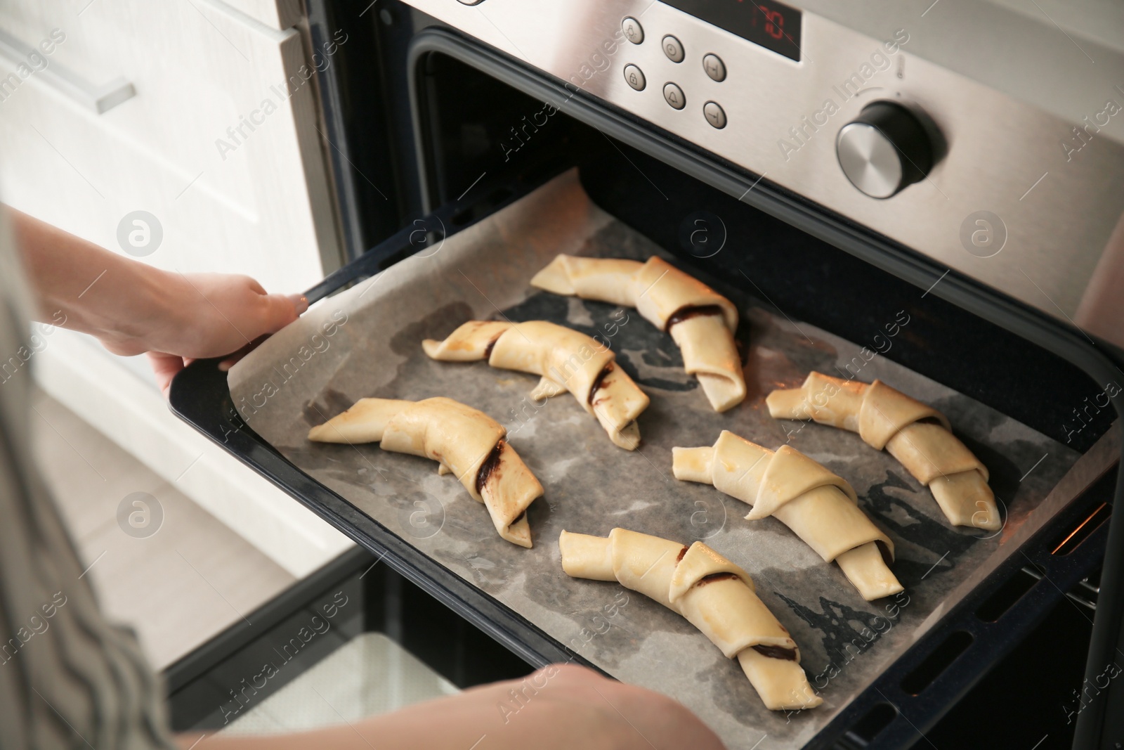 Photo of Woman putting baking sheet with raw croissants in oven, closeup