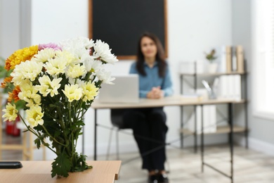 Photo of Pedagogue at table in classroom, focus on bouquet. Teacher's day