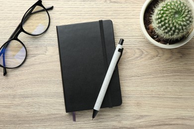 Closed black notebook, pen, glasses and cactus on wooden table, flat lay