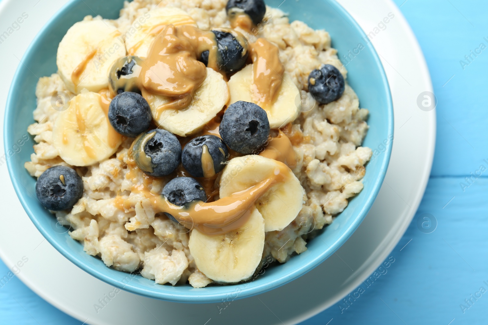 Photo of Tasty oatmeal with banana, blueberries and peanut butter served in bowl on light blue wooden table, closeup