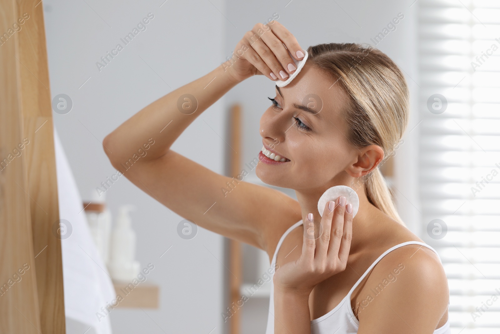 Photo of Smiling woman removing makeup with cotton pads in bathroom