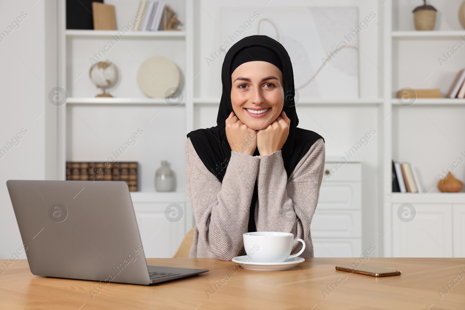 Photo of Muslim woman with cup of drink near laptop at wooden table in room