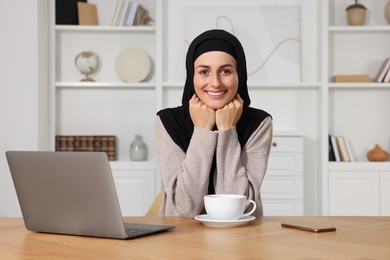 Muslim woman with cup of drink near laptop at wooden table in room