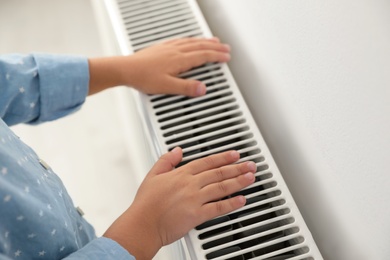 Photo of Child warming hands on heating radiator near white wall, closeup