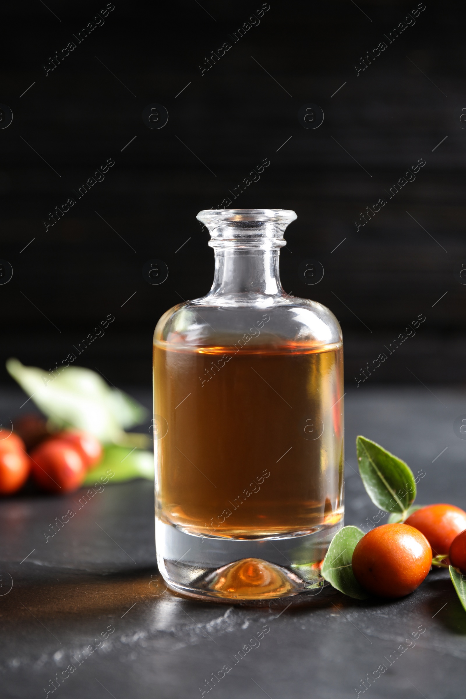 Photo of Glass bottle with jojoba oil and seeds on grey stone table against dark background