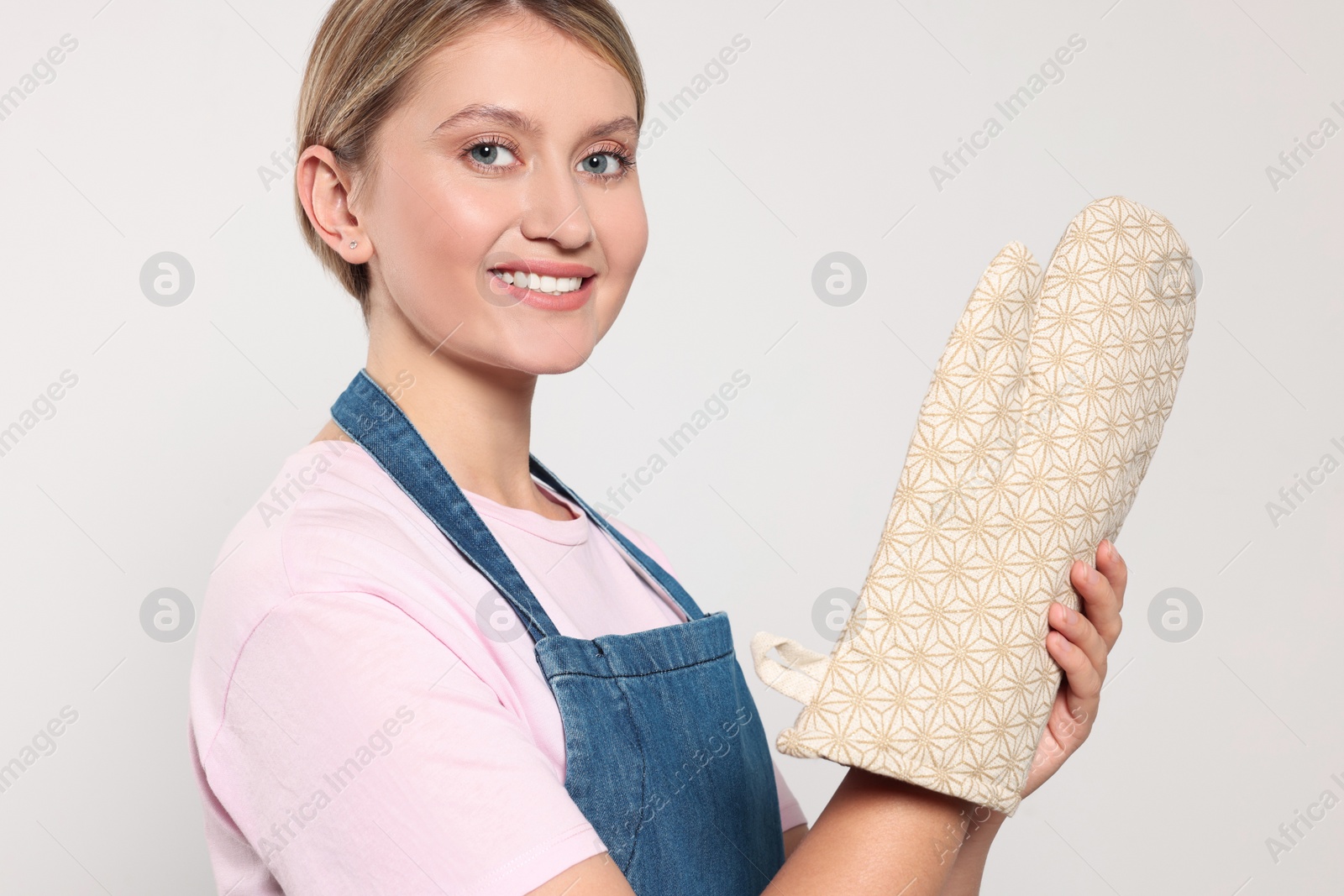 Photo of Beautiful young woman in denim apron and oven glove on light grey background, closeup