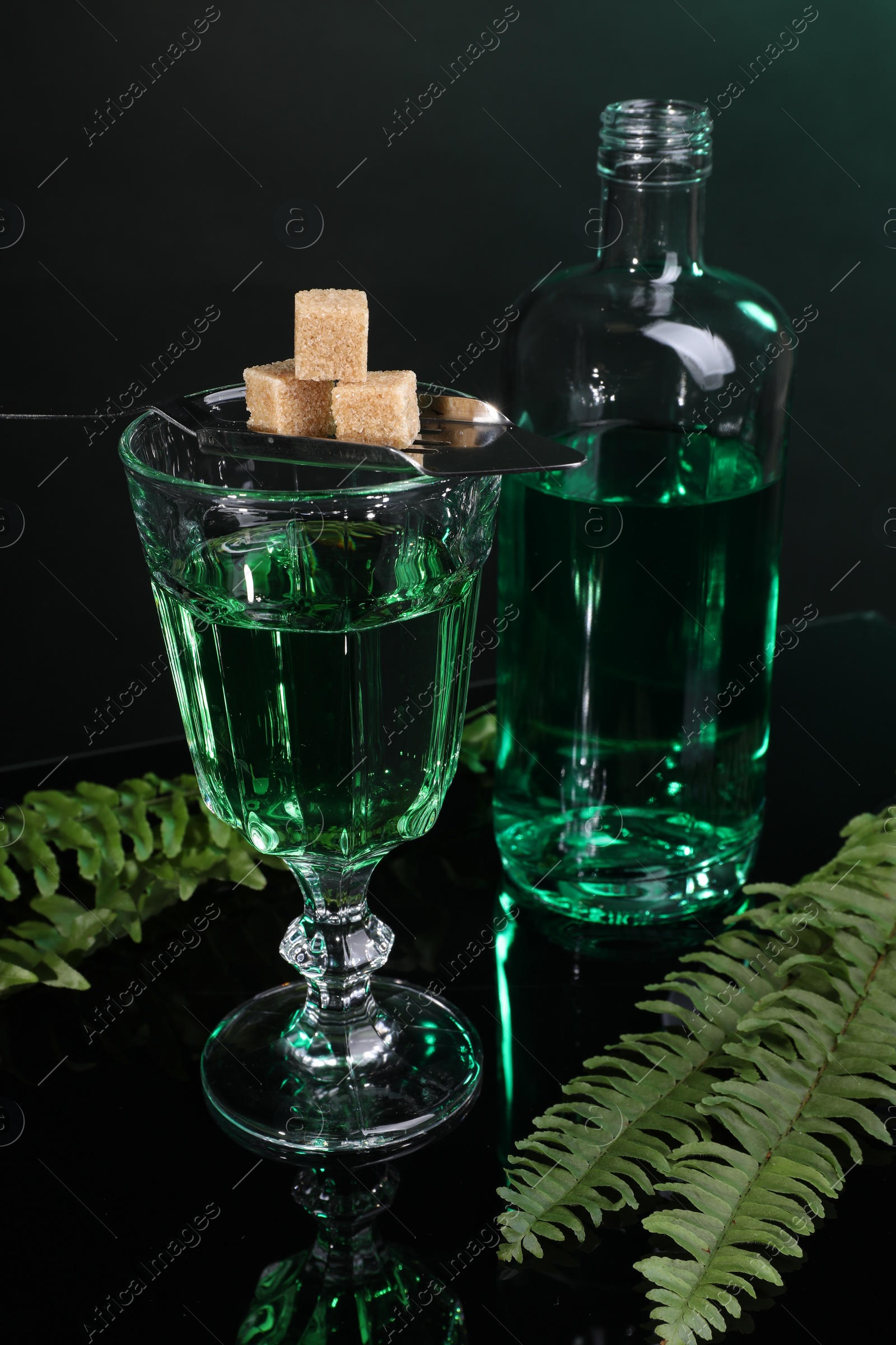 Photo of Absinthe, brown sugar, spoon and green leaves on mirror table. Alcoholic drink