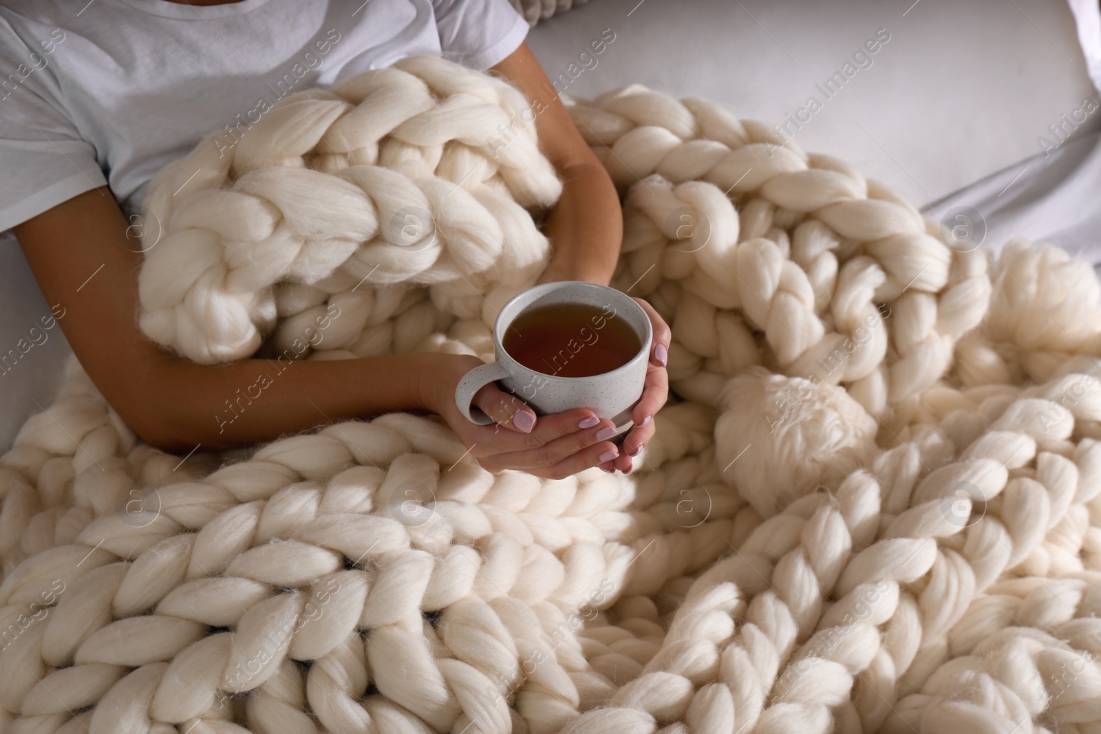 Photo of Woman covered with knitted plaid holding cup of tea in bed, closeup