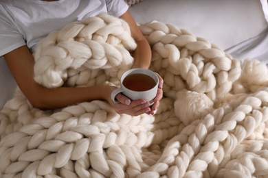 Woman covered with knitted plaid holding cup of tea in bed, closeup