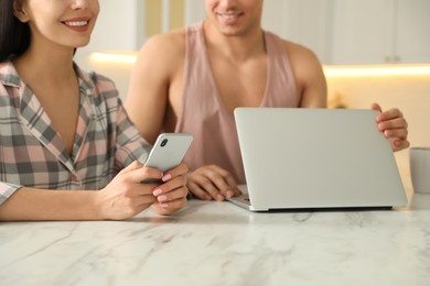 Photo of Happy couple wearing pyjamas with gadgets spending time together in kitchen, closeup