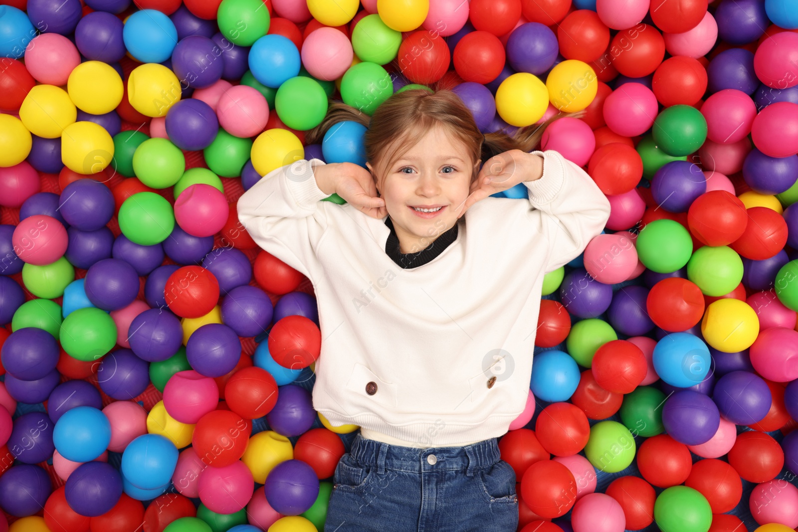 Photo of Happy little girl lying on many colorful balls, top view