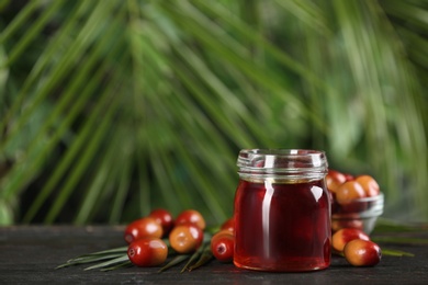 Photo of Palm oil in glass jar, tropical leaf and fruits on wooden table. Space for text