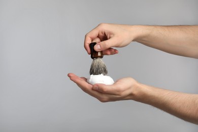 Man applying shaving foam onto brush on light grey background, closeup