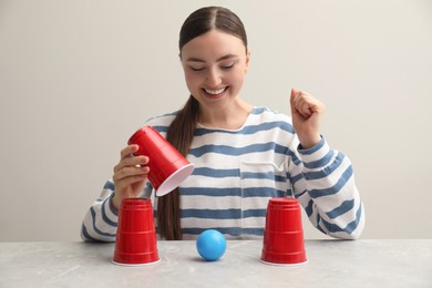 Photo of Shell game. Happy woman showing ball under cup at light marble table
