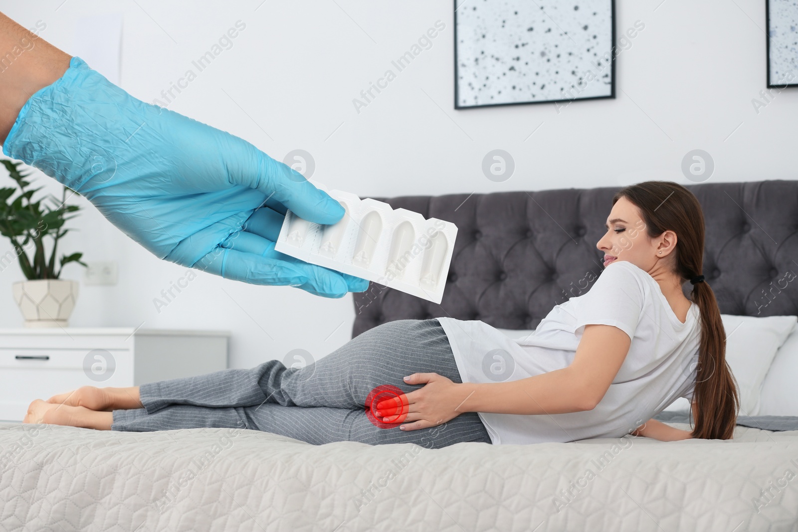 Image of Doctor holding suppositories for hemorrhoid treatment and woman suffering from pain on bed at home