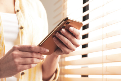 Young woman using modern smartphone near window indoors, closeup