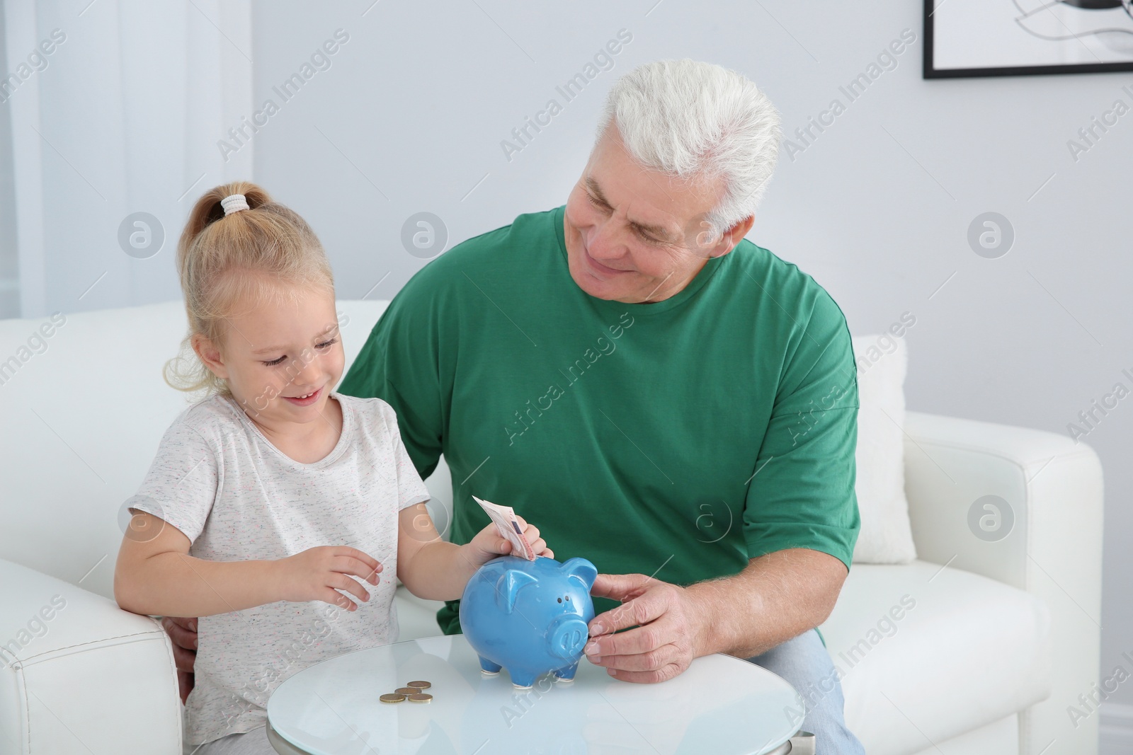 Photo of Little girl putting money into piggy bank and her grandfather at table