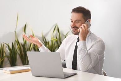 Happy man using modern laptop while talking on smartphone at desk in office