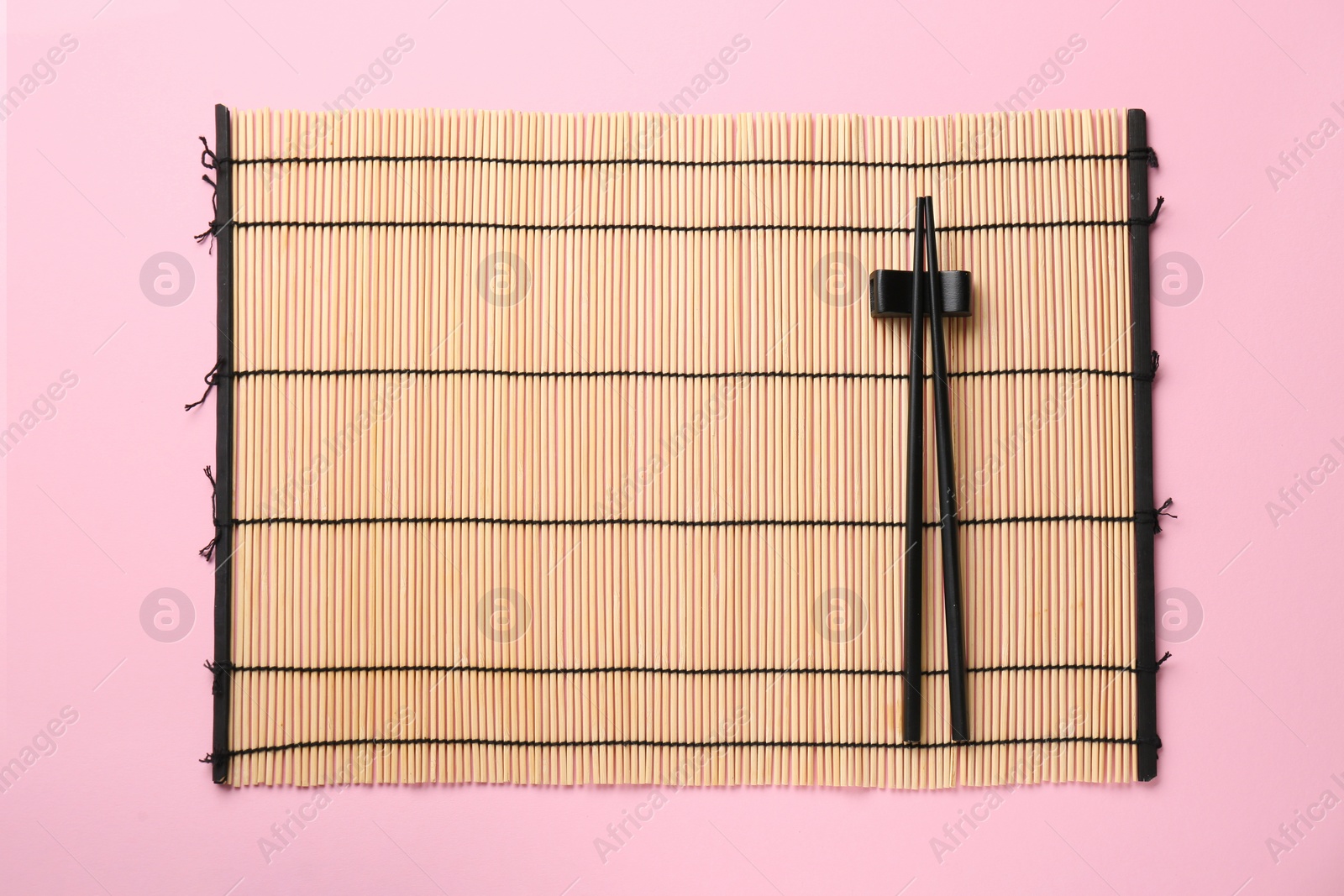 Photo of Bamboo mat with pair of black chopsticks and rest on pink background, top view